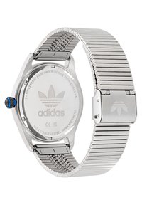 Nerūsējošā tērauda pulkstenis ar sudraba acu sietu joslu, astoņstūra korpusu, zilu kroni un iegravētu Adidas logotipu aizmugurē un sprādzes daļā.