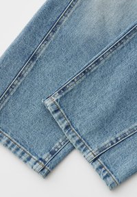 Gros plan sur les jambes d'un jean en denim bleu clair montrant les coutures détaillées et les ourlets pliés sur un fond blanc.