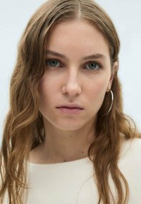 Gros plan sur un visage avec des cheveux bouclés brun. Le sujet a les yeux verts, un maquillage minimal et porte une boucle d'oreille en argent circulaire. Vêtements de couleur neutre.