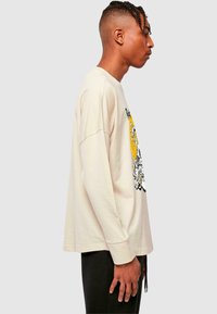 Beige Langarm-T-Shirt aus Baumwolle mit einem grafischen Druck in Gelb- und Schwarztönen. Lockere Passform mit gerippten Bündchen.