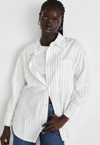 Donna con capelli intrecciati indossa una camicia bianca a righe oversized sopra una maglietta bianca e jeans blu, in piedi contro un muro bianco uniforme.