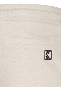 Tissu beige clair avec une poche rectangulaire à l'arrière et un logo noir carré dans le coin inférieur droit. Texture douce.
