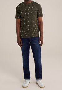 Camiseta de manga corta verde olivo con patrón disperso blanco, combinada con jeans azul oscuro y zapatillas blancas con suelas de goma.