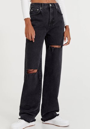 Jean noir taille haute à jambes larges avec déchirures aux genoux, associé à des baskets blanches et un top court blanc à manches longues.