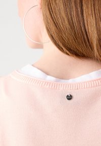 Personne aux cheveux roux portant un pull rose pâle sur une chemise blanche, visible de dos avec une grande boucle d'oreille argentée et une étiquette sur le pull.