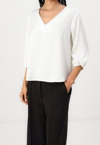 Blouse blanche à col en V avec manches bouffantes trois-quarts ; tissu lisse, coupe décontractée. Associée à un pantalon noir ample avec poches.