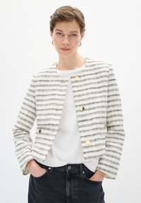 Skrzacowany, pleciony żakiet w kremowym i czarnym kolorze, z złotymi guzikami, skróconym krojem i okrągłym dekoltem. Stylizowany z białą koszulą i czarnymi dżinsami.