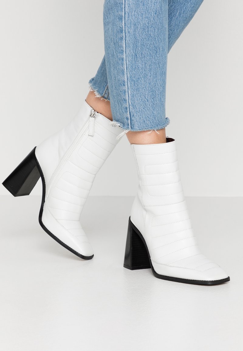 Femme portant des bottines blanches à talons hauts avec des talons carrés noirs et un jean à ourlet effiloché bleu clair, posant devant un fond uni.