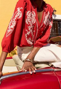 Röd blus med vit blommig broderi, V-ringning, puffärmar och elastiska manschetter. Metallarmband på handen. Ljust färgade byxor, vintagebil i bakgrunden.