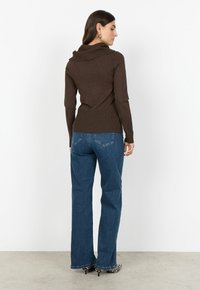 Kvinna vänd bort från kameran, iklädd en brun lång�ärmad tröja, blå raka jeans och klackar med svart-vitt mönster.