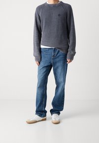 Person in einem grauen Strickpullover, blauen Jeans und weißen Sneakers, der vor einem schlichten hellen Hintergrund steht.