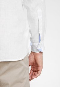 Biała lniana koszula z jasnoniebieskimi akcentami przy mankietach. Posiada dopasowany fason, długie rękawy oraz zapięcie na guziki przy mankietach.