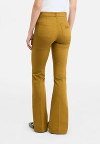 Eng anliegende, ausgestellte senfgelbe Hosen mit hoher Taille, Gesäßtaschen und einem Logopatch auf der rechten Seite über der Tasche.