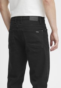 Black denim jeans with a straight cut, featuring a leather logo patch on the waistband, two back pockets, and contrast stitching details.