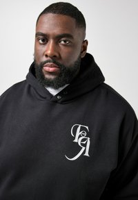 Primer plano de un hombre con barba que lleva una sudadera con capucha negra con un logotipo bordado en blanco en el pecho, sobre un fondo claro y liso.