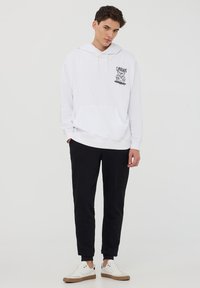 Felpa bianca con tasca frontale e un design grafico nero. Abbinata a pantaloni neri e scarpe bianche con accenti beige.