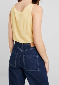 Gelbes ärmelloses Oberteil mit tief ausgeschnittenem Rücken, kombiniert mit dunkelblauen, hoch Taillierten Jeanshosen mit zwei Gesäßtaschen und sichtbaren Nähten.