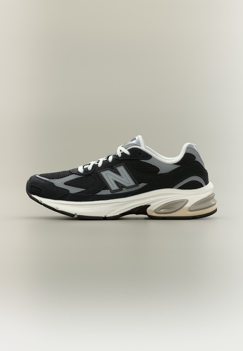Chaussure de course New Balance noire et grise avec des lacets blancs et une semelle blanche épaisse, vue de profil sur un fond neutre.