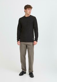 Schwarzer gerippter Pullover mit Rundhalsausschnitt, kombiniert mit hellgrauen Hosen und schwarzen Schuhen, vor einem hellen Hintergrund stehend.
