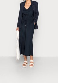 Tmavě modré culotte šaty s elastickým pasem, doplněné o odpovídající blazer. Bílá platformová sandály dotvářejí vzhled. Drží pruhovanou tašku.