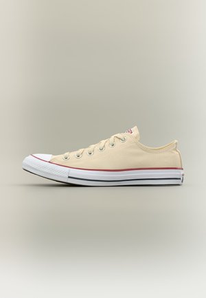 Zapatilla beige de lona baja con puntera y suela de goma blanca, detalles de rayas rojas y negras y cordones blancos, mostrada de lado.