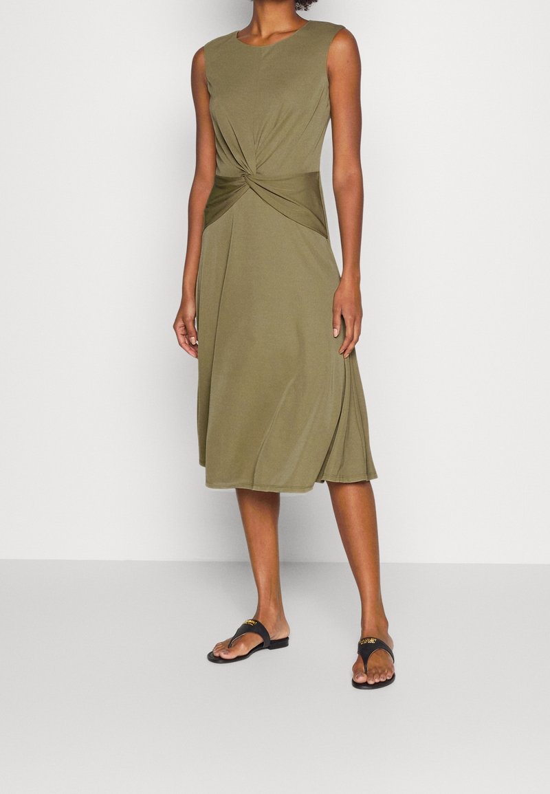 Sukienka bez rękawów w oliwkowym kolorze z przewiązanym węzłem w talii, zwiewny fason w kształcie litery A oraz miękka, gładka tekstura. Założona z czarnymi sandałami.