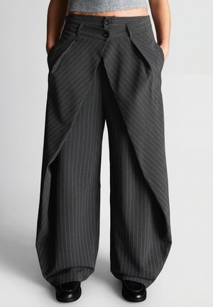 Žena nosiaca vysoké pásové, široké nohavice s pruhmi, prekryté predné panely a ruky v vreckách, v kombinácii s čiernymi topánkami.