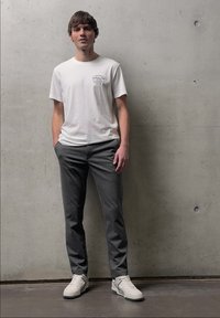 Jeune homme debout contre un mur en béton gris portant un t-shirt blanc imprimé, un pantalon gris foncé et des baskets blanches.