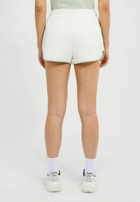 Persona que lleva puestos pantalones cortos blancos con ribete verde claro, calcetines blancos y zapatillas blancas, de pie contra un fondo claro y liso.