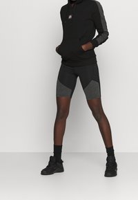 Sweat à capuche noir avec accents gris. Shorts noirs ajustés avec panneaux gris clair. Portés avec des chaussures de sport noires et des chaussettes aux chevilles.
