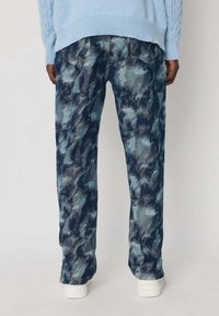 Personne portant un pantalon ample à motif camouflage bleu et gris avec des poches arrière, un pull léger bleu clair et des chaussures blanches, se tenant de dos.
