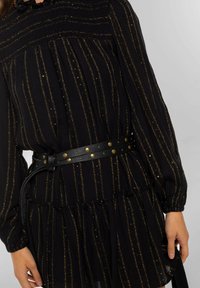 Vestido negro con rayas verticales doradas, tejido texturizado, escote redondo y mangas largas abullonadas. Cinturón negro con tachuelas doradas ciñe la cintura.