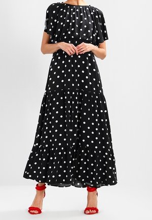 Frau trägt ein schwarzes, kurzärmeliges, gestuftes Maxikleid mit weißen Punkten und roten High-Heel-Sandalen.