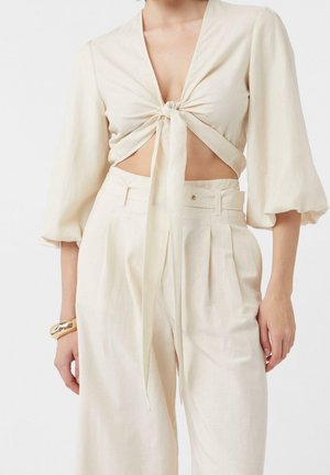 Kvinde iført cremefarvet cropped bluse med knude foran og matchende plisserede bukser med høj talje, med et gyldent armbånd om venstre håndled.