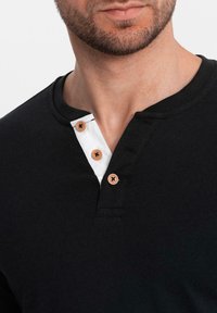 Zwarte katoenen henley-shirt met een witte placket en drie bruine knopen. Zachte textuur met een geribde kraag. Close-upweergave.
