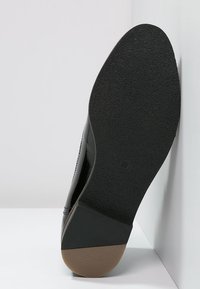 Zapato negro con un acabado brillante, que presenta una suela texturizada, punta redondeada y un leve tacón. La suela es de goma oscura con un agarre estampado.