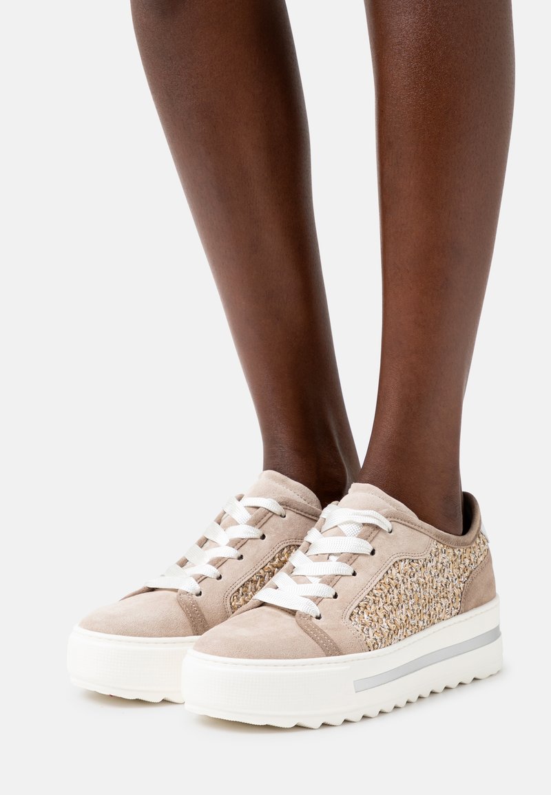Baskets à plateforme beige avec lacets blancs et panneaux latéraux tissés portées sur des jambes à la peau foncée sur fond blanc.