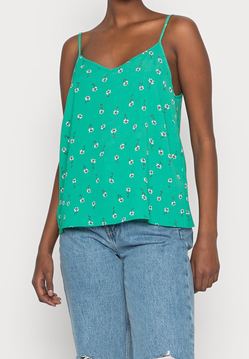 Haut caraco vert avec un décolleté en V, fines bretelles et petit motif floral blanc, assorti à un short en jean bleu clair déchiré.