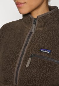 Sudadera de forro polar marrón con cuello alto, cremallera frontal y un pequeño bolsillo en el pecho. Presenta una superficie texturizada y suave, además de un parche con logo.