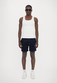 Débardeur blanc, shorts bleu marine et baskets blanches. Le modèle se tient droit, porte des lunettes de soleil noires. Tissu lisse et ajusté avec un design simple.