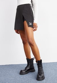 Jupe-short de sport noire avec une coupe mi-haute et un design fendu. Fabriquée en tissu texturé avec un logo Puma blanc sur le côté. Bottes de combat noires.