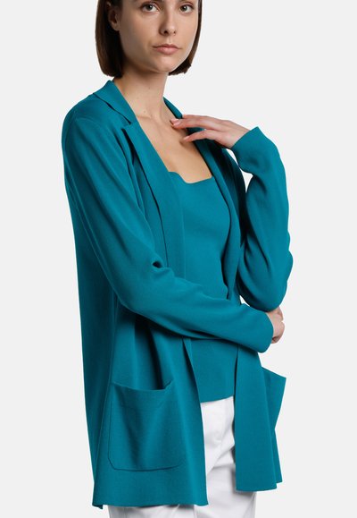 Cardigan turquoise avec une coupe décontractée, à l'avant ouvert, manches longues et deux poches avant. Texture lisse, tissu léger. Superposé à un haut assorti.