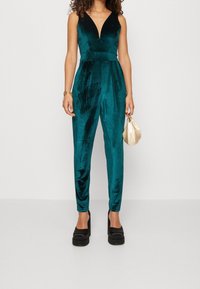 Teal sammetsbyxdress med djupt V-ringning, figurnära liv och smala ben. Bärs med svarta platåskor och håller en crèmefärgad clutchtvätt.