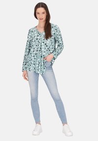 Een lichtgroene blouse met zwart-witte stippen, met een V-hals, lange mouwen en een relaxte pasvorm, gecombineerd met lichtblauwe jeans en witte sneakers.