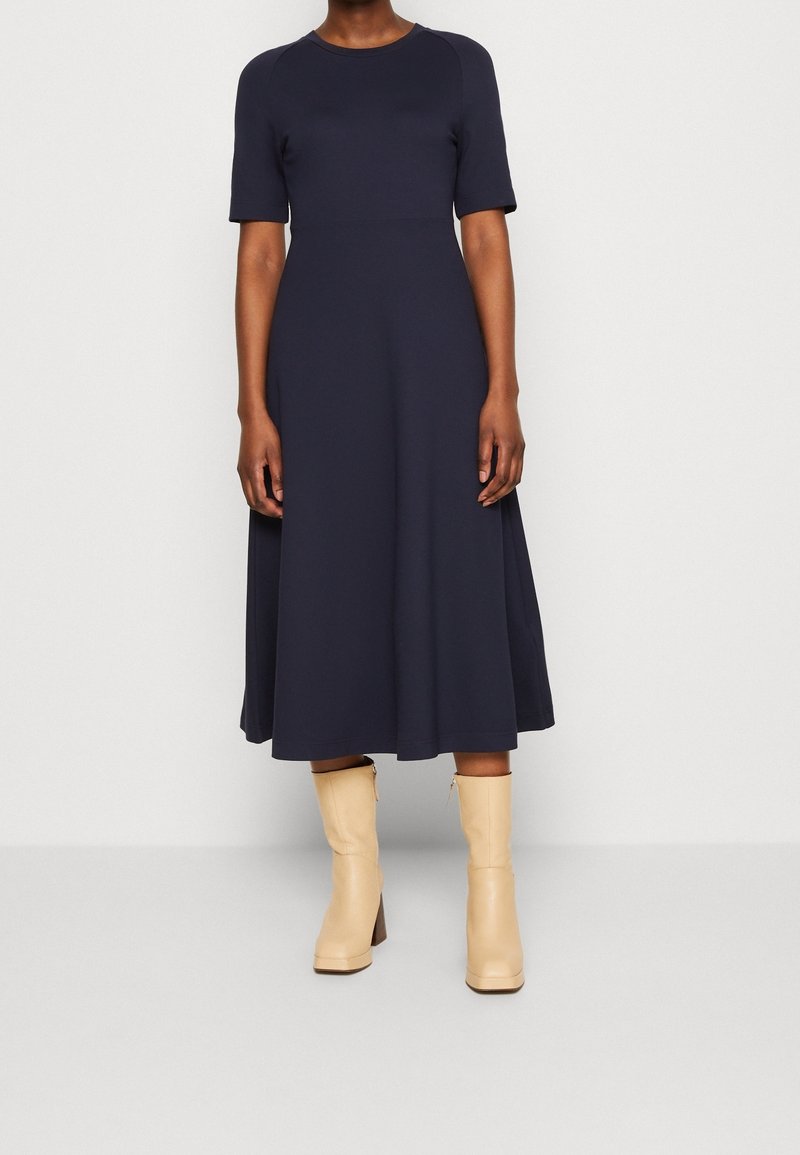 Robe bleu marine longueur genoux avec manches courtes, en tissu lisse. Modèle portant des bottes beiges montantes jusqu'aux genoux avec talons carrés.