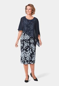 Marineblaues Blumenkleid mit Chiffonüberzug und weißen Blumenmustern, knielang, kombiniert mit schwarzen Absätzen. Glatte Textur und elegantes Design.