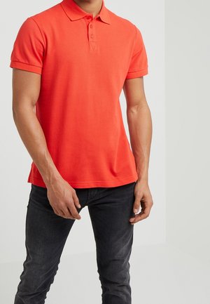 Homme portant un polo à manches courtes orange vif et un jean gris foncé, debout, les bras détendus le long du corps, devant un fond uni.