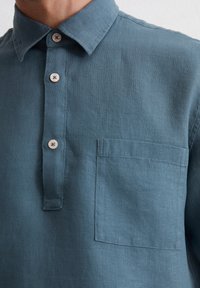 Button-up shirt made of lightweight blue linen, featuring a pointed collar, three buttons, and a single chest pocket.