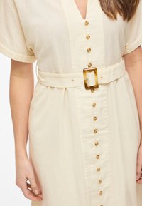 Robe crème à manches courtes boutonnée avec une boucle de ceinture rectangulaire en écaille cintrant la taille.