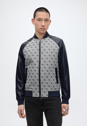 Jeune homme portant une veste zippée avec un devant gris à motifs et des manches en cuir noirs, debout devant un fond clair uni.